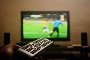 Stream4k Close-up of a hand holding a remote control in front of a TV showing a football match.